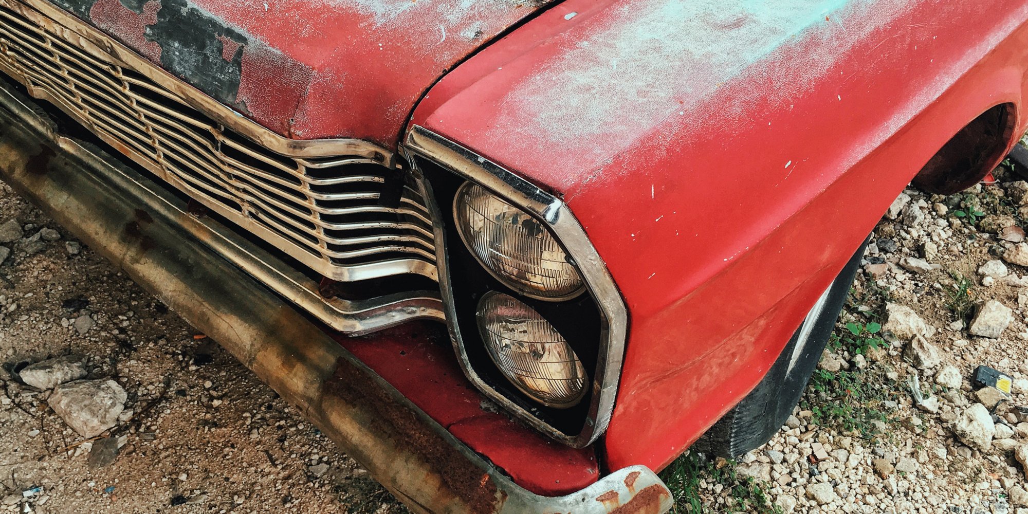 Detailaufnahme eines alten, verrosteten roten Autos mit verblasstem Lack und beschädigter Stoßstange.