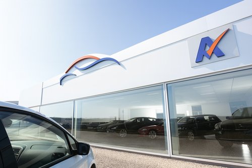 Helles Foto eines modernen Autohauses mit großer Glasfront; links im Vordergrund die Front eines weißen Pkw, dahinter im Showroom mehrere Fahrzeuge in einer sonnigen, klaren Tages­szene.