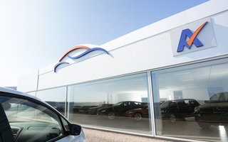 Helles Foto eines modernen Autohauses mit großer Glasfront; links im Vordergrund die Front eines weißen Pkw, dahinter im Showroom mehrere Fahrzeuge in einer sonnigen, klaren Tages­szene.