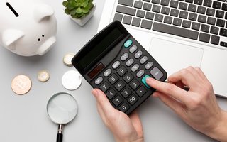 Hände tippen auf einem Taschenrechner neben Laptop, Sparschwein und Münzen auf einem Schreibtisch.