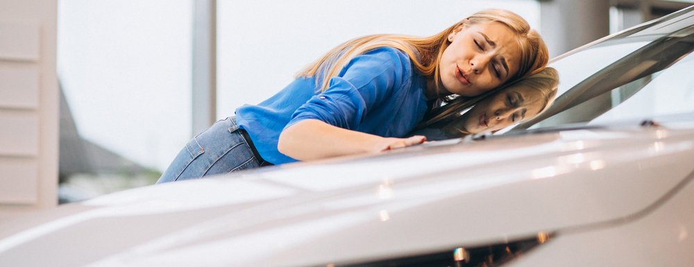 Junge Frau lehnt sich glücklich an ein Auto und streicht über die Motorhaube.