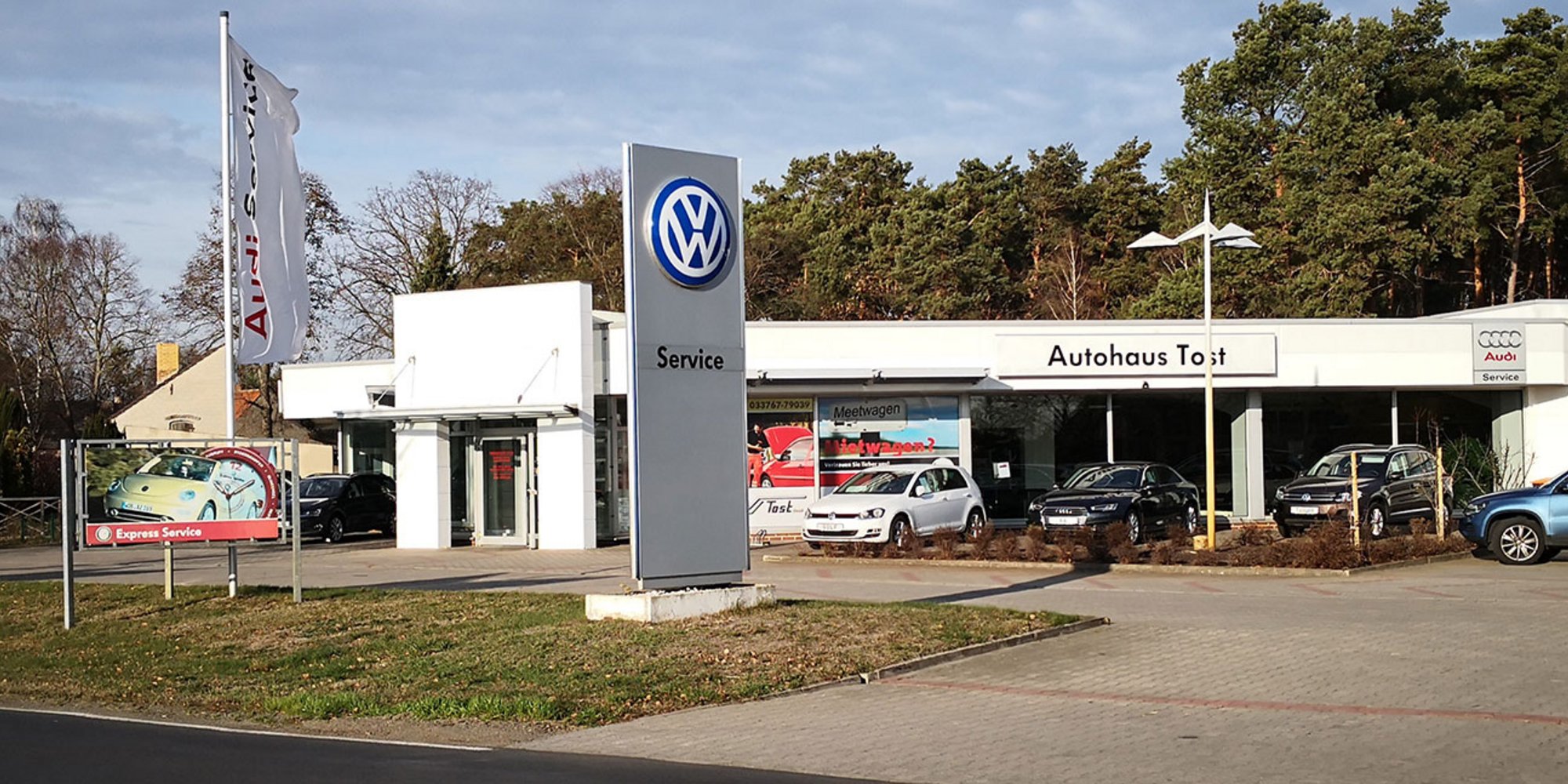ußenansicht eines Autohauses mit VW-Logo, mehreren Fahrzeugen davor und Bäumen im Hintergrund. 