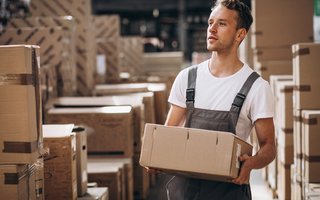Arbeiter trägt einen Karton durch ein Lager mit vielen gestapelten Versandkartons.