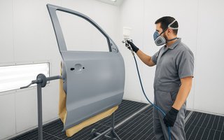 Ein Mann trägt Schutzmaske und Handschuhe und lackiert eine Autotür in einer Lackierkabine mit einer Spritzpistole.