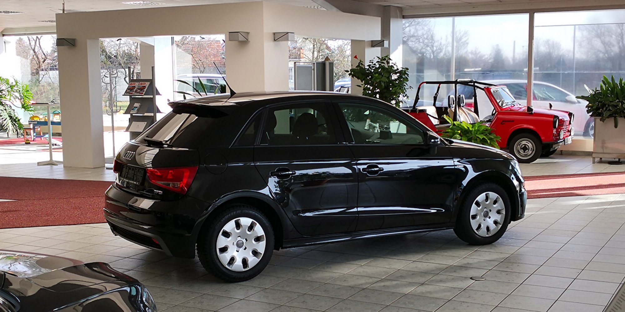 Innenansicht eines Autohauses mit schwarzem Kleinwagen im Vordergrund und roten Teppichwegen.